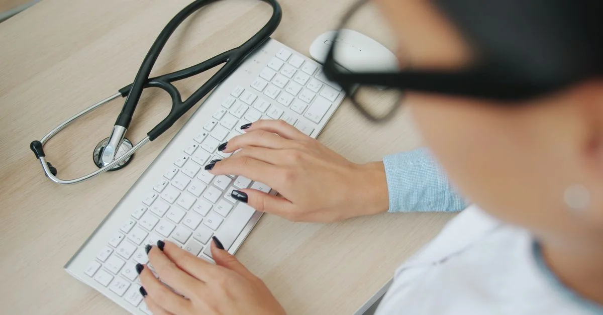 Le médecin utilise un clavier d'ordinateur ; un stéthoscope se trouve à proximité, sur le bureau.