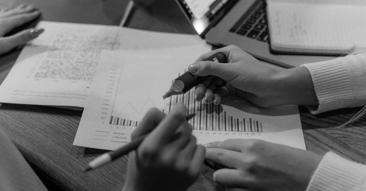 Image monochrome de mains analysant des documents et des graphiques dans un environnement de bureau.